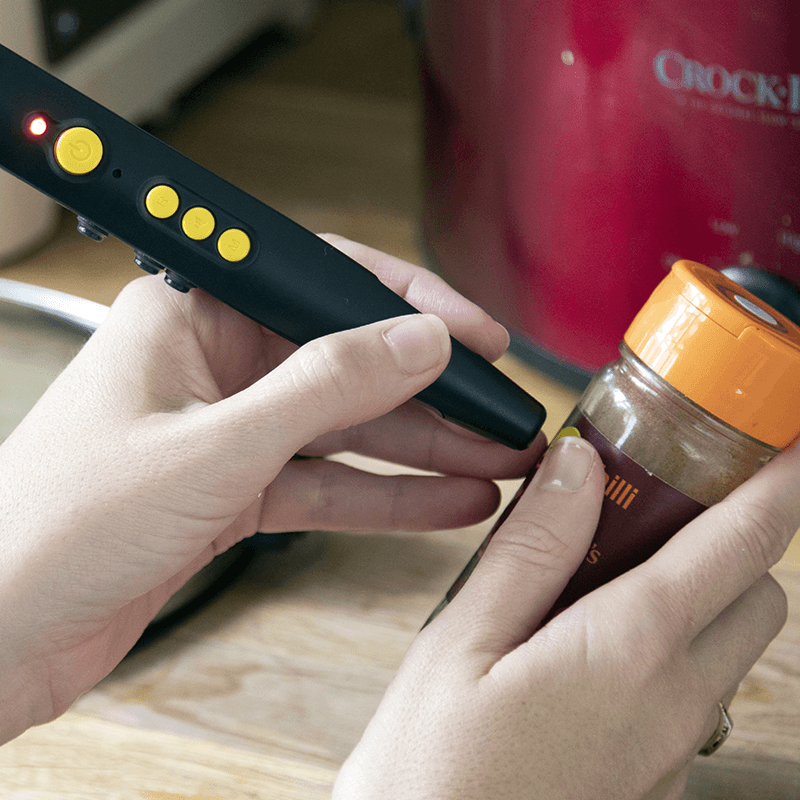 Na zdjęciu widoczne są dłonie trzymające niewielkie urządzenie w kształcie długopisu oraz mały słoiczek z przyprawą. Urządzenie jest czarne, z kilkoma żółtymi, wypukłymi przyciskami i świecącą diodą. Końcówka urządzenia skierowana jest w stronę etykiety na słoiczku, co sugeruje skanowanie lub odczyt informacji. Słoiczek ma przezroczyste ścianki i pomarańczową zakrętkę. Scena przedstawia korzystanie z urządzenia do głosowego etykietowania produktów, które umożliwia osobom niewidomym rozpoznawanie przedmiotów poprzez nagrane komunikaty dźwiękowe.