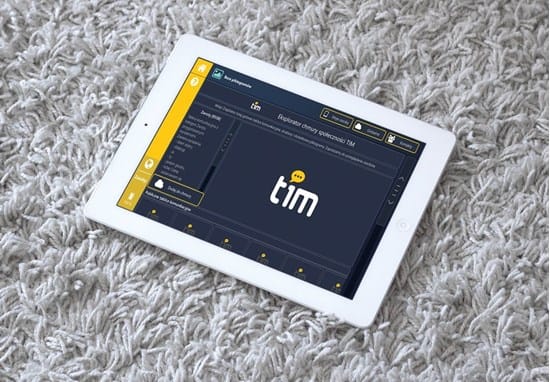 Na zdjęciu widoczny jest tablet leżący na jasnym, miękkim dywanie. Na ekranie uruchomiona jest aplikacja do komunikacji alternatywnej. Interfejs aplikacji ma ciemne tło z żółtymi elementami menu po bokach. W centralnej części ekranu wyświetlone jest słowo „tim” z ikoną dymku rozmowy, sugerującą komunikację. Po lewej stronie widoczne jest menu z kategoriami lub opcjami, a na górze pasek narzędzi. Urządzenie służy do wspierania komunikacji i edukacji osób mających trudności w porozumiewaniu się.