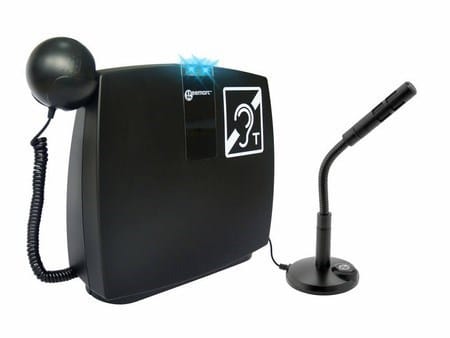 Na zdjęciu widoczny jest zestaw do wspomagania słyszenia – przenośna pętla indukcyjna. Składa się z czarnego, prostokątnego urządzenia głównego z symbolem ucha oraz litery „T”, oznaczającym kompatybilność z aparatami słuchowymi. Po lewej stronie podłączona jest słuchawka telefoniczna na spiralnym przewodzie, a po prawej mikrofon na elastycznym ramieniu ustawiony na podstawce. Zestaw służy do poprawy jakości dźwięku dla osób korzystających z aparatów słuchowych, szczególnie w rozmowach i sytuacjach wymagających komunikacji.