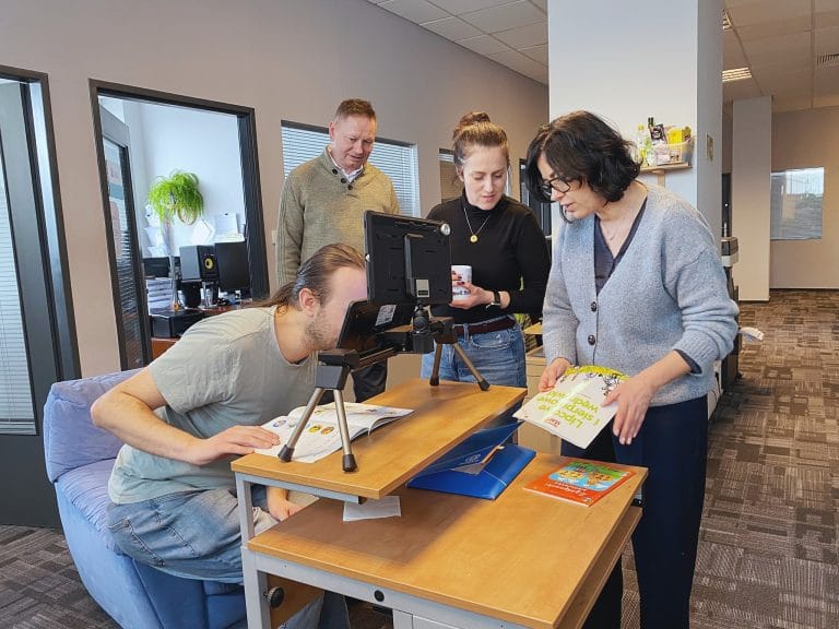 Na zdjęciu widoczne są cztery osoby w przestrzeni biurowej, pracujące wspólnie przy stole. Na pierwszym planie mężczyzna siedzi i pochyla się nad urządzeniem powiększającym ustawionym na statywie, oglądając książkę lub dokument. Obok stoją trzy osoby – dwie kobiety i mężczyzna – które obserwują działanie sprzętu. Jedna z kobiet trzyma otwartą książkę, druga patrzy na urządzenie, a mężczyzna stoi nieco z tyłu. Na stole znajdują się książki, dokumenty oraz laptop. W tle widać biuro z przeszklonymi pomieszczeniami i rośliną. Sytuacja przedstawia testowanie technologii wspierającej czytanie i pracę z tekstem.