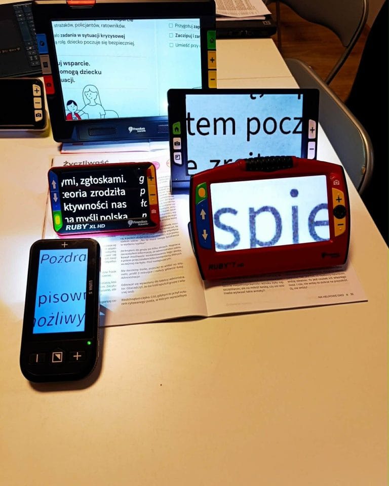 Na zdjęciu widocznych jest kilka elektronicznych powiększalników ustawionych na stole, służących do odczytywania tekstu przez osoby słabowidzące. Urządzenia mają różne kształty i kolory – czarne, czerwone i kolorowe – i wyposażone są w przyciski do regulacji powiększenia oraz kontrastu. Każdy z powiększalników wyświetla powiększony fragment tekstu z otwartej książki lub dokumentu leżącego pod nimi. Na ekranach widać duże, wyraźne litery o różnym kontraście (np. jasne litery na ciemnym tle lub odwrotnie). W tle znajduje się większy ekran również pokazujący powiększony tekst. Całość przedstawia zestaw technologii wspierających czytanie i dostęp do informacji dla osób z niepełnosprawnością wzroku.