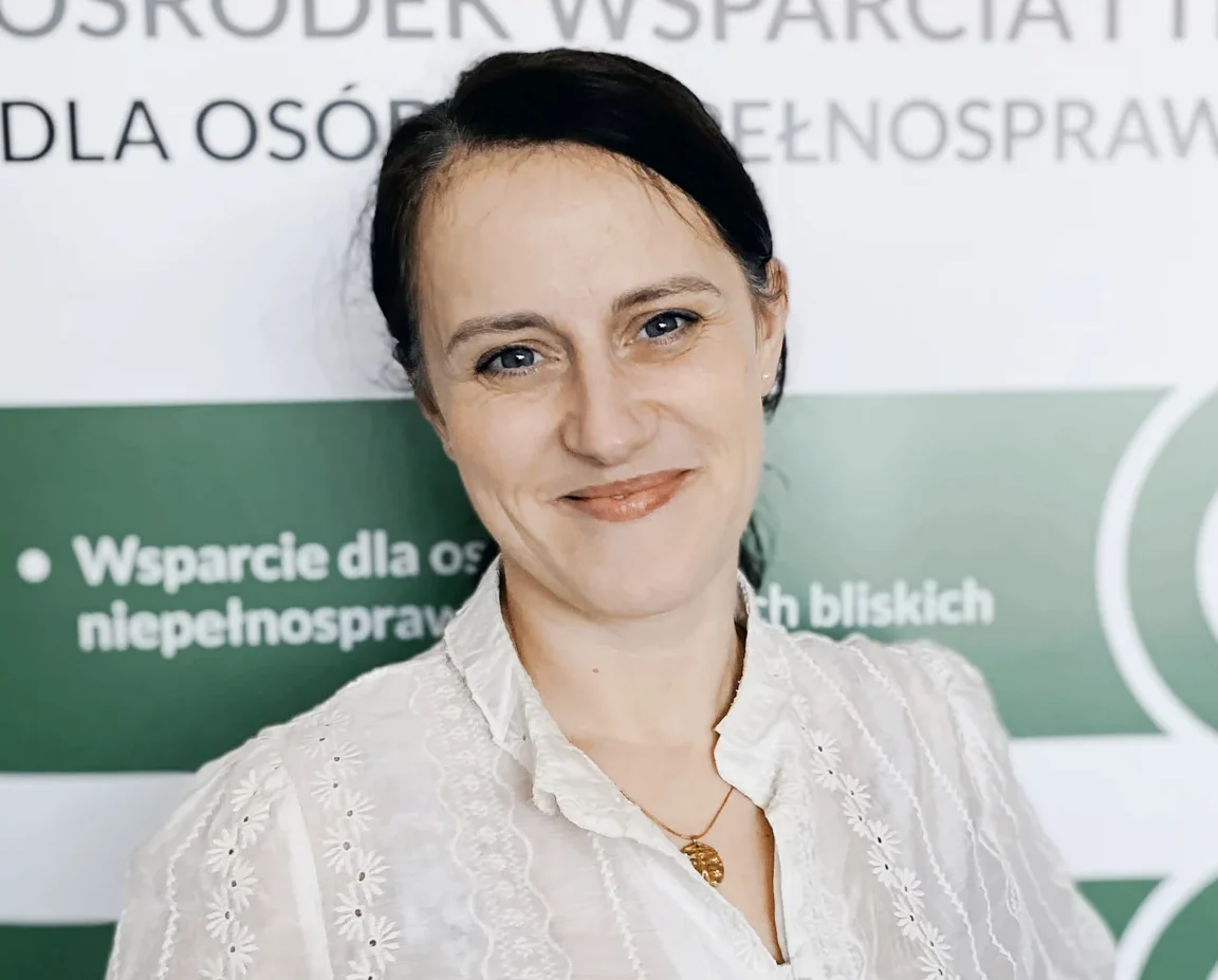 Agnieszka Sobolewska-Czokajlo Na zdjęciu widoczna jest kobieta stojąca na tle roll-upu informacyjnego dotyczącego wsparcia dla osób z niepełnosprawnościami. Kobieta ma ciemne włosy związane do tyłu i ubrana jest w jasną, białą bluzkę z delikatnym wzorem. Ma na szyi cienki łańcuszek z zawieszką. Patrzy w stronę aparatu i uśmiecha się. W tle znajduje się baner z fragmentami tekstu w języku polskim, w tym informacjami o wsparciu dla osób z niepełnosprawnościami i ich bliskich. Całość ma charakter profesjonalny i promocyjny.