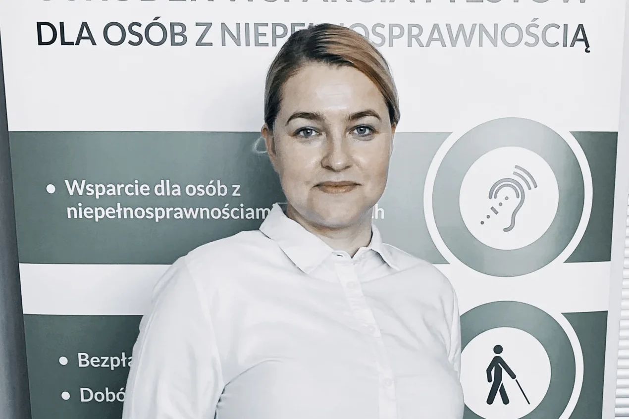 Aleksandra Szalek Palmowska Na zdjęciu widoczna jest kobieta stojąca na tle roll-upu informacyjnego dotyczącego wsparcia dla osób z niepełnosprawnościami. Kobieta ma jasne włosy związane do tyłu i ubrana jest w białą koszulę. Patrzy w stronę aparatu z lekkim uśmiechem. W tle znajduje się baner z napisami w języku polskim, m.in. „dla osób z niepełnosprawnością” oraz punktami dotyczącymi wsparcia. Po prawej stronie widoczne są piktogramy, m.in. symbol ucha (dotyczący słuchu) oraz postaci z białą laską (osoba niewidoma). Całość ma charakter informacyjno-promocyjny.