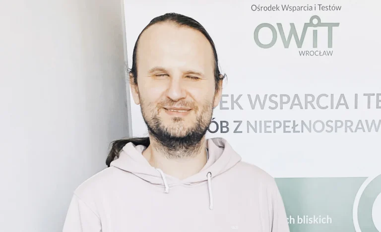 Witold Strugała Na zdjęciu widoczny jest mężczyzna stojący na tle roll-upu informacyjnego Ośrodka Wsparcia i Testów (OWiT Wrocław). Mężczyzna ma długie, ciemne włosy związane z tyłu oraz krótki zarost. Ubrany jest w jasną bluzę z kapturem. Patrzy w stronę aparatu i lekko się uśmiecha. W tle znajduje się baner z logo „OWiT Wrocław” oraz fragmentami tekstu dotyczącego wsparcia dla osób z niepełnosprawnościami. Całość ma charakter profesjonalny i informacyjny.