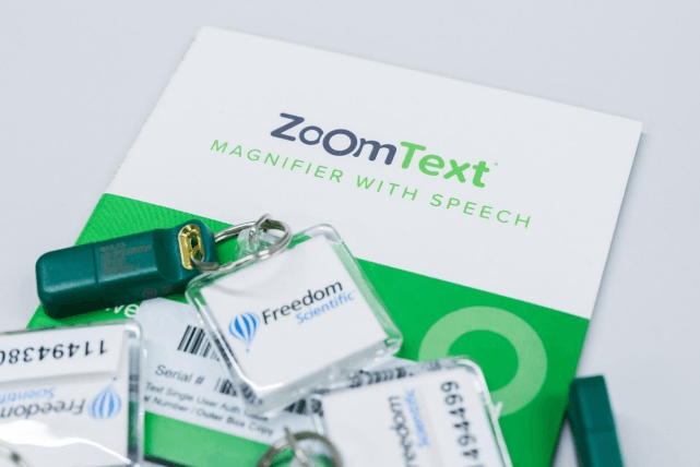 Na zdjęciu widoczne są materiały związane z oprogramowaniem „ZoomText Magnifier with Speech”. W tle znajduje się ulotka lub opakowanie w kolorach białym i zielonym z nazwą programu. Na pierwszym planie leżą breloki i pendrive’y z logo Freedom Scientific oraz etykietami licencyjnymi, zawierającymi numery seryjne. Całość przedstawia zestaw oprogramowania powiększającego z funkcją mowy, przeznaczonego dla osób słabowidzących do powiększania obrazu i odczytu tekstu na komputerze.