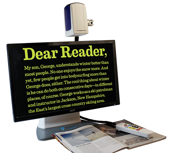 Na zdjęciu widoczny jest stacjonarny powiększalnik elektroniczny do czytania. Składa się z monitora ustawionego na podstawie oraz kamery zamocowanej nad ekranem. Na ekranie wyświetlony jest powiększony tekst w wysokim kontraście – żółte litery na czarnym tle, zaczynający się od słów „Dear Reader”. Pod monitorem znajduje się otwarta książka, którą kamera powiększa. Obok leży pilot z przyciskami do sterowania urządzeniem. Sprzęt służy osobom słabowidzącym do wygodnego czytania tekstu w powiększeniu.