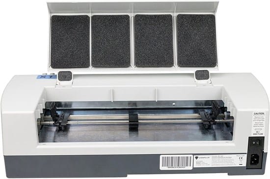 Na zdjęciu widoczna jest drukarka brajlowska z otwartą pokrywą. Urządzenie ma prostokątną, jasnoszarą obudowę. W górnej części widać podniesioną klapę z czterema panelami wygłuszającymi. Wewnątrz znajduje się mechanizm drukujący z metalową prowadnicą i elementami przesuwającymi papier. Z przodu widoczny jest otwór na arkusze, a po prawej stronie panel z przyciskiem zasilania i gniazdem. Urządzenie służy do drukowania tekstów w alfabecie Braille’a dla osób niewidomych.